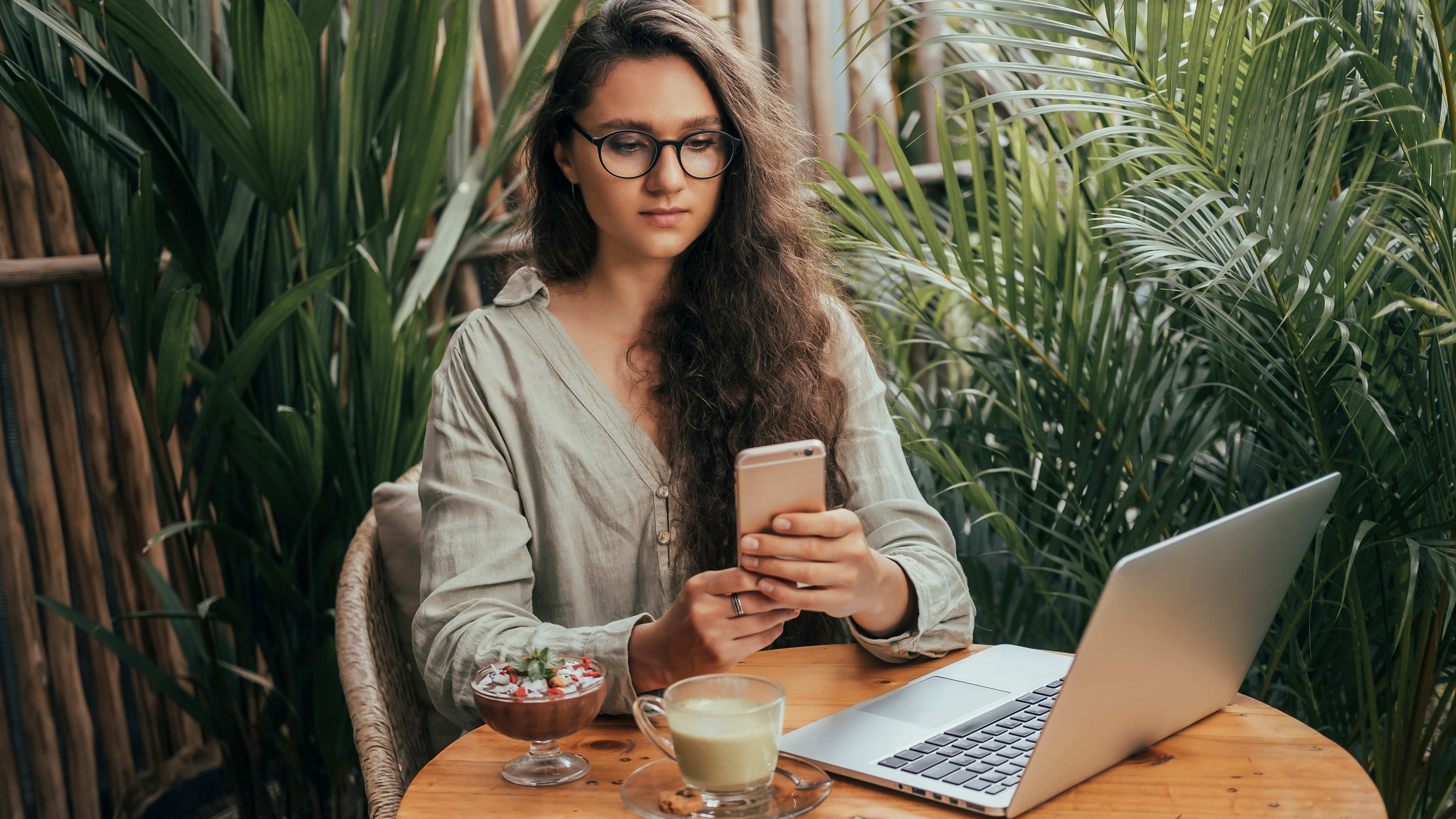 A woman sitting at a table and holding an iPhone, and she has a laptop open on the table