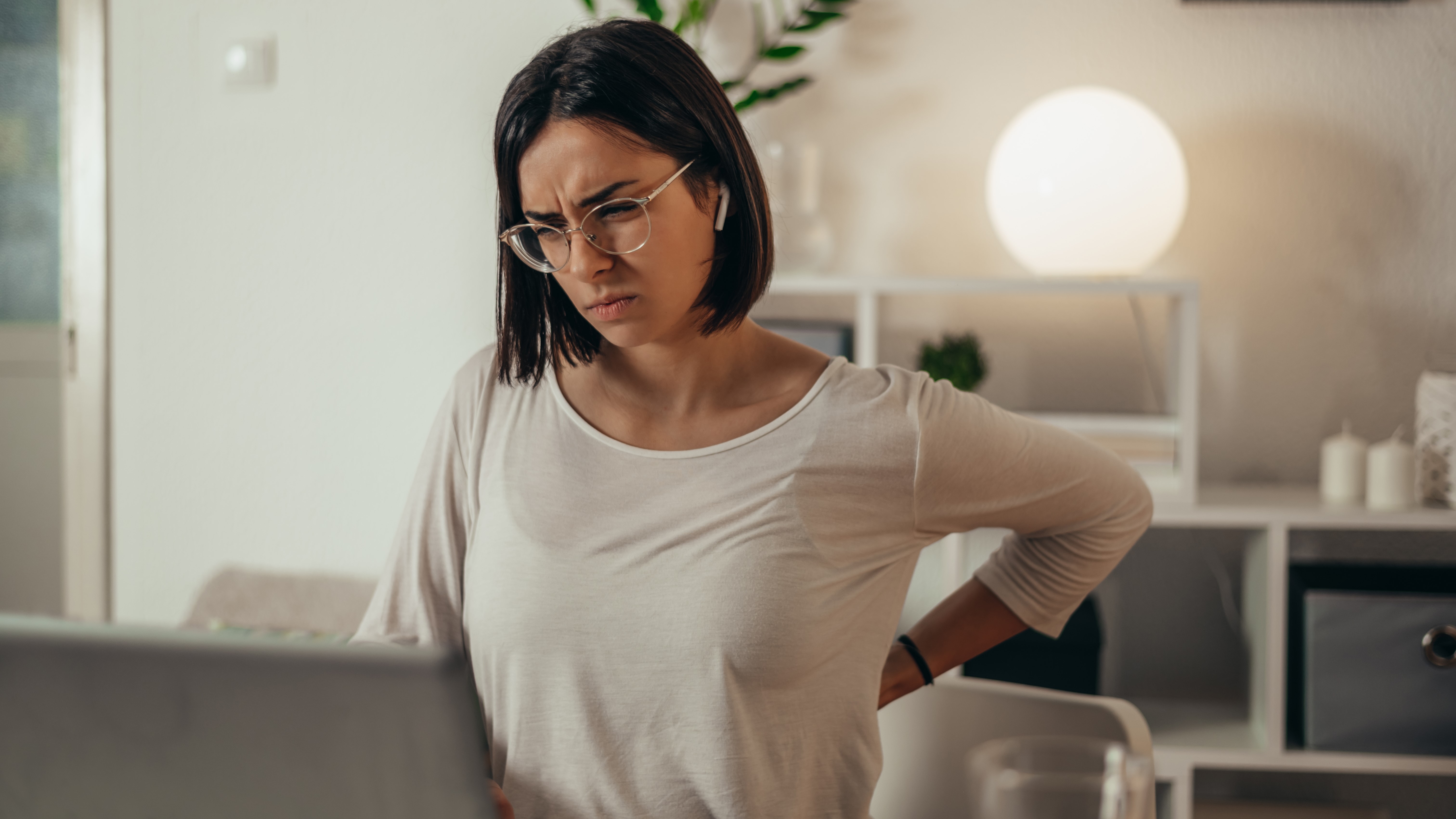 A woman arching holding her back with one arm and looking at a laptop situated on a surface